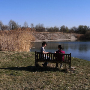 Fernes Foto von einer Bank, auf der Rese und N. sitzen und beratend miteinander sprechen. Im Hintergund See, Schilf, Ufer und blauer Himmel.